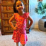 child, girl, portrait, standing, smiling, dress, floral_pattern, coral_color, hands_on_hips, indoors, home, bedroom, dresser, wooden_furniture, houseplant, framed_photos, carpet, fashion, full_body, family_photo