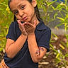 child, portrait, outdoor, smiling, playful, blowing_kiss, face, eyes, hair, hair_accessory, hands, arms, shirt, plaid_clothing, short_sleeves, tattoo, greenery, plants, garden, bokeh