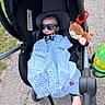baby, stroller, sunglasses, blanket, plush_toy, outdoor, pavement, hand, greenery, toy, person, infant, clothing, sunlight, footwear, parent, accessory, shade, cute, relaxation