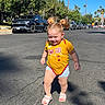 child, toddler, girl, smiling, outdoor, street, sunny, daylight, yellow_shirt, sports_shirt, iowa_state, shorts, sandals, pigtails, standing, walking, parked_cars, pickup_truck, palm_trees, residential_neighborhood
