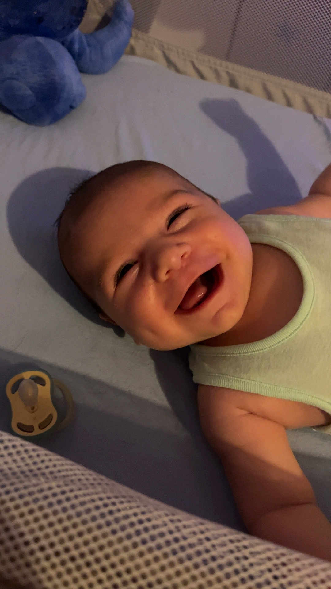 Liam participe au concours pour gagner de l'argent avec cette photo : baby, smiling, infant, lying_down, pacifier, plush_toy, elephant_toy, crib, soft_surface, tank_top, happy, cute, indoors, shadow, child, face, expression, skin, clothing, nursery