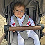 baby, stroller, child, infant, outdoor, seatbelt, smiling, blue_eyes, white_clothing, heart_pattern, socks, face, person, pavement, handlebar, casual, cute, young_child, portrait, daylight