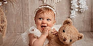 Lyna a rejoint le concours — aidez-le/la à gagner de superbes lots ! baby, child, teddy_bear, stuffed_animal, white_dress, headband, smiling, happy, indoor, wooden_floor, wooden_wall, decor, flowers, soft_toy, cute, portrait, playful, infant, close_up, cozy