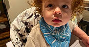James is registered to the contest to win money with this photo: toddler, child, high_chair, curly_hair, blue_bib, indoor, wooden_floor, face, person, clothing, baby, furniture, table, plant, curious, expression, portrait, young_child, sitting, home
