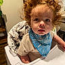 toddler, child, high_chair, curly_hair, blue_bib, indoor, wooden_floor, face, person, clothing, baby, furniture, table, plant, curious, expression, portrait, young_child, sitting, home