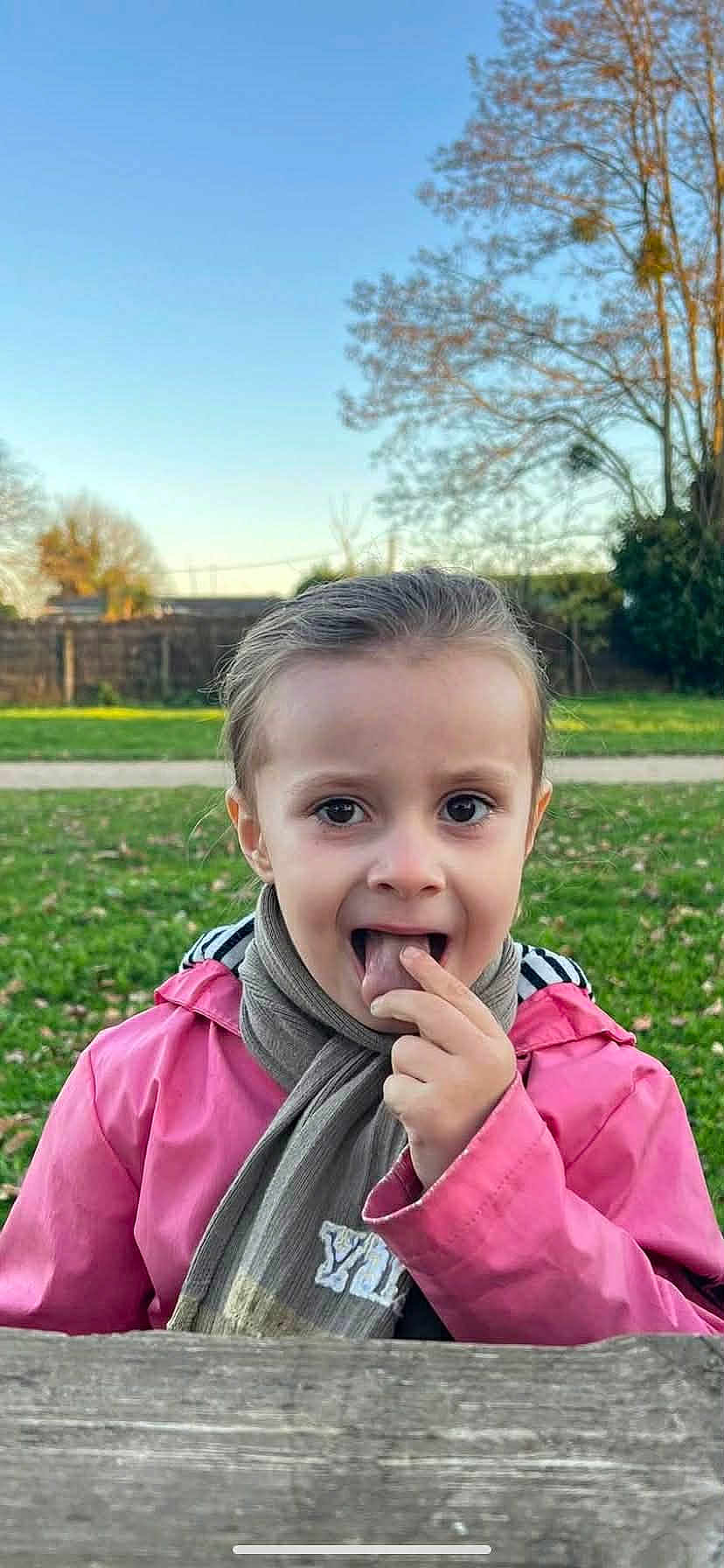 Helena Catalia a rejoint le concours — aidez-le/la à gagner de superbes lots ! child, pink_jacket, scarf, outdoor, park, grass, tree, wooden_table, sky, fall, playful, face, person, young, nature, daylight, casual_clothing, expression, curious, portrait