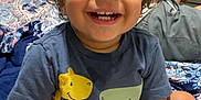 Jamari is registered to the contest to win money with this photo: child, toddler, smiling, curly_hair, bed, bedding, cross_legged, blue_shirt, shorts, indoor, happy, person, young_child, cute, portrait, sitting, room, furniture, drawer, casual