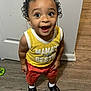 toddler, child, curly_hair, yellow_shirt, red_shorts, black_shoes, white_socks, wooden_floor, door, wall, happy, smiling, standing, indoors, person, excited, eyes_wide_open, clothing, flooring, expression