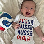Liam joined the competition — help win amazing prizes! baby, infant, smiling, onesie, text_on_clothing, soccer_ball, lying_down, blanket, happy, child, cute, indoor, face, person, blue_eyes, short_hair, white_clothing, patterned_blanket, playful, joyful