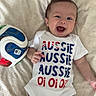 baby, infant, smiling, onesie, text_on_clothing, soccer_ball, lying_down, blanket, happy, child, cute, indoor, face, person, blue_eyes, short_hair, white_clothing, patterned_blanket, playful, joyful