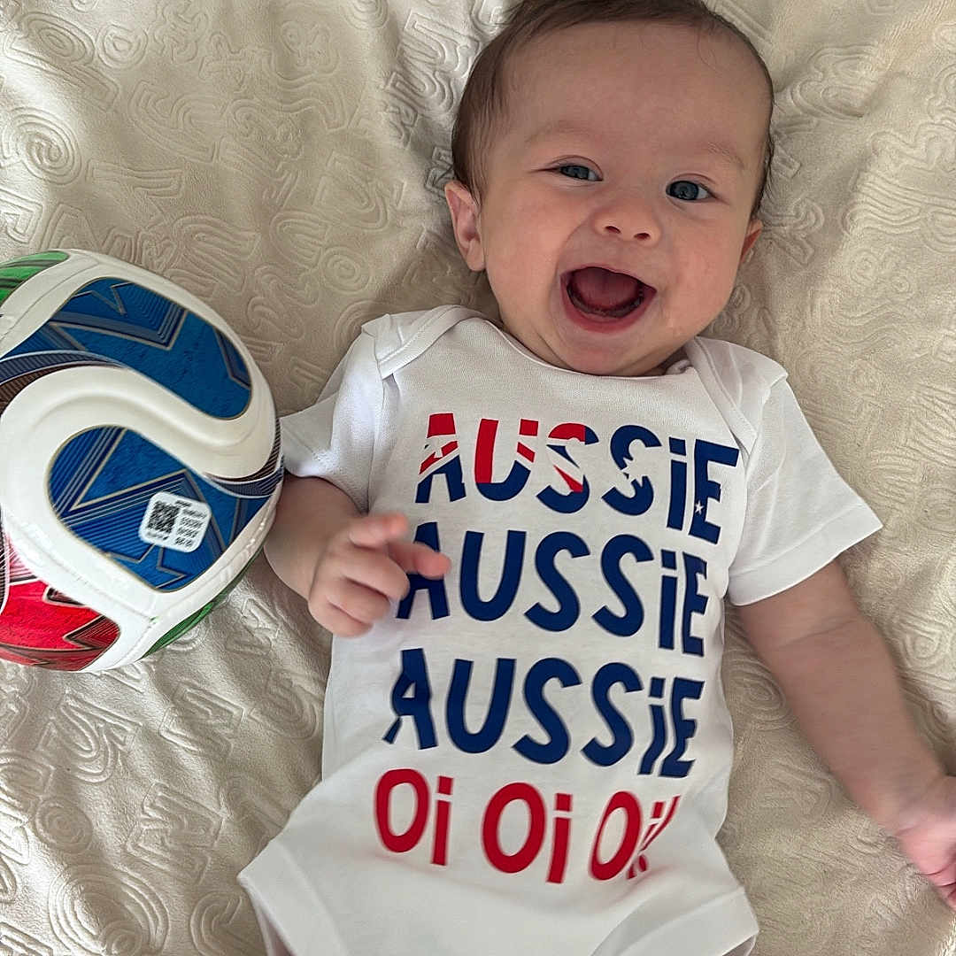 Liam joined the competition — help win amazing prizes! baby, infant, smiling, onesie, text_on_clothing, soccer_ball, lying_down, blanket, happy, child, cute, indoor, face, person, blue_eyes, short_hair, white_clothing, patterned_blanket, playful, joyful