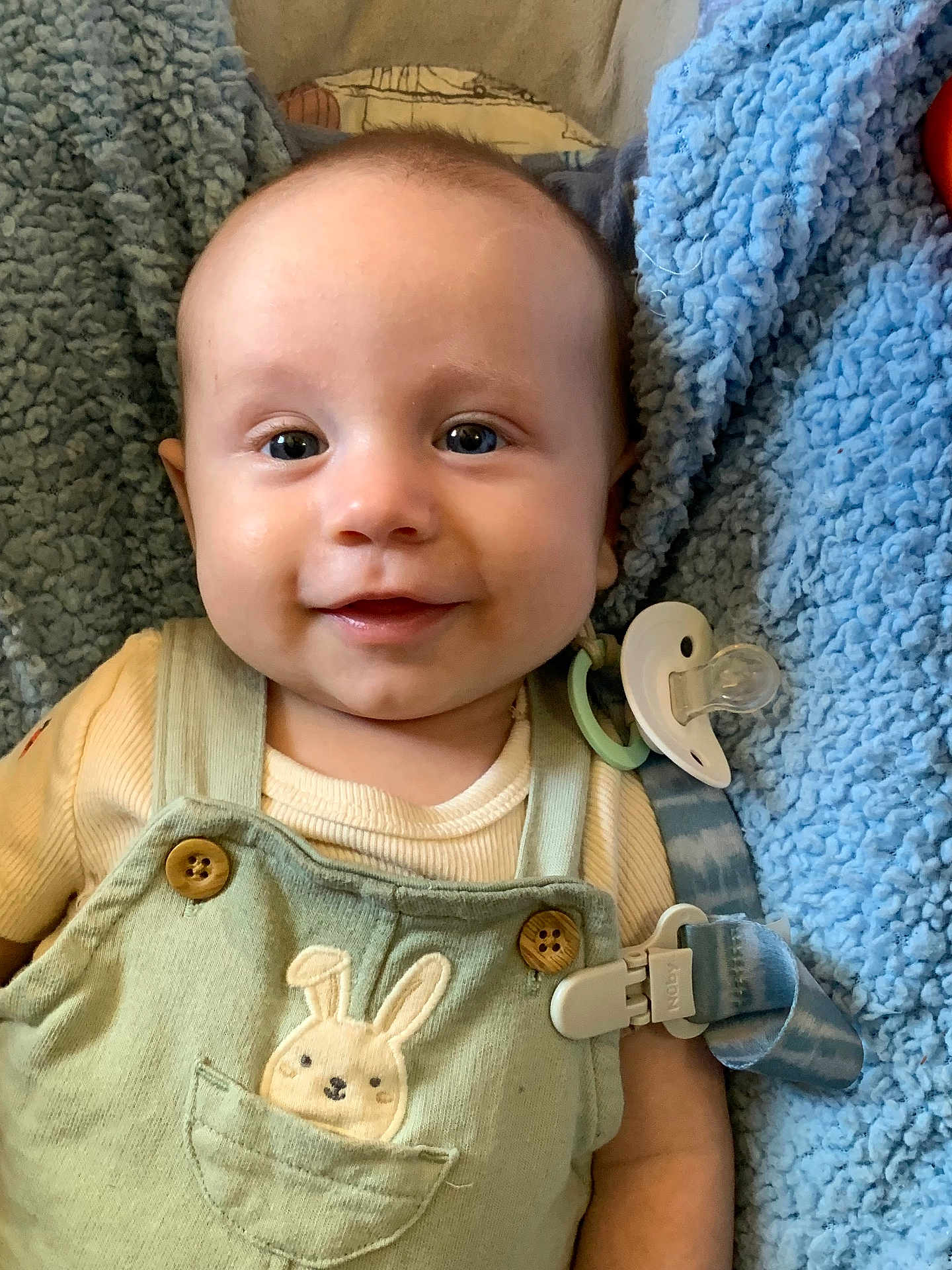 Oliver joined the competition — help win amazing prizes! baby, smiling, infant, overalls, bunny, pacifier, blanket, blue, clothing, cute, happy, face, child, lying_down, soft, comfort, indoors, closeup, button, person