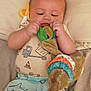 baby, child, toy, plush, shirt, pants, shark_pattern, truck_pattern, bed, blanket, infant, cute, soft_toy, lying_down, indoors, playful, colorful, hands, face, focus