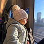 toddler, child, winter_clothing, knit_hat, coat, window, train, sunlight, profile, urban, blurred_background, indoor, transportation, warm_light, face, person, travel, looking_out, soft_texture, hand