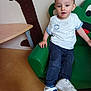 casual_clothing, child, curious_expression, dark_pants, floor, frog_shape, furniture, green_chair, indoor, person, playroom, sitting, table, toddler, tree_decal, wall_decal, white_shirt, white_shoes, yellow_object, young_child