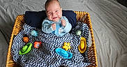 Paxton is registered to the contest to win money with this photo: baby, basket, blanket, blue_clothing, chevron, child, colorful, cute, face, happy, indoor, infant, pattern, person, playful, portrait, smile, toy, toys, wicker