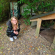 Kylliana participe au concours pour gagner de l'argent avec cette photo : child, glasses, black_jacket, outdoor, gravel, greenery, tree, wooden_structure, playhouse, leaves, crouching, smiling, nature, casual_clothing, daylight, person, young_girl, red_glasses, shoes, adventure