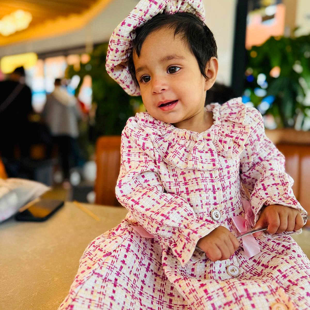 Divyanka Mahajan is registered to the contest to win money with this photo: blurry_background, buttoned_dress, child, curious, cute, dress, fashion, hat, holding_object, indoor, long_sleeves, patterned_clothing, person, portrait, seated, smiling, striped_socks, table, toddler, young_child