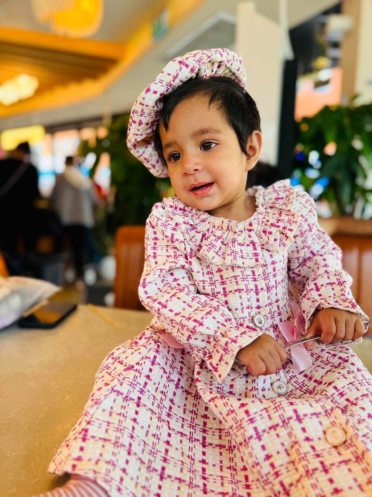 Divyanka Mahajan is registered to the contest to win money with this photo: toddler, child, dress, hat, table, indoor, person, cute, patterned_clothing, holding_object, curious, portrait, smiling, blurry_background, seated, fashion, young_child, long_sleeves, buttoned_dress, striped_socks