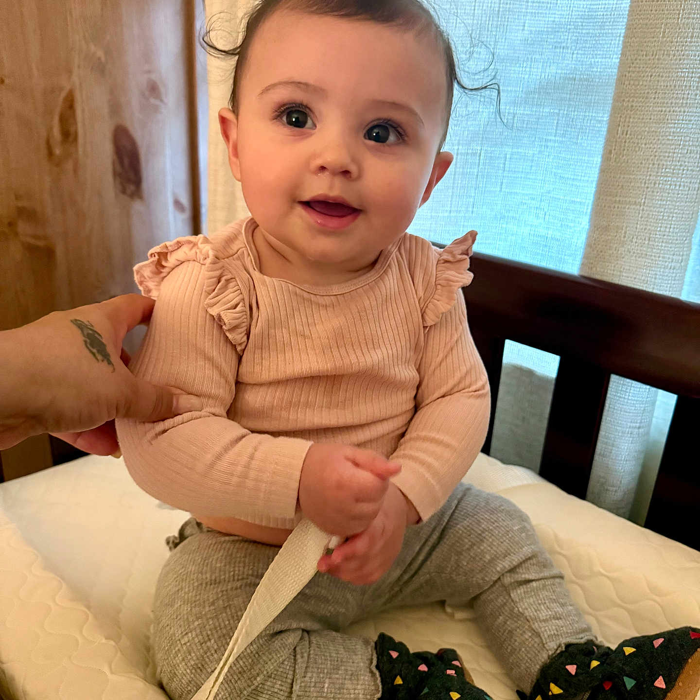 Sydney is registered to the contest to win money with this photo: baby, booties, bright_eyes, crib, curly_hair, curtain, hand, indoor, infant, leggings, long_sleeve, mattress, onesie, portrait, rosy_cheeks, sitting, smiling, strap, tattoo_on_hand, wooden_furniture