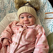 Winsley is registered to the contest to win money with this photo: baby, child, hat, knit_hat, pom_poms, plaid_jacket, pink_clothing, baby_seat, cushion, warning_label, indoor, smiling, face, hand, comfortable, portrait, cozy, apparel, person, warm_clothing