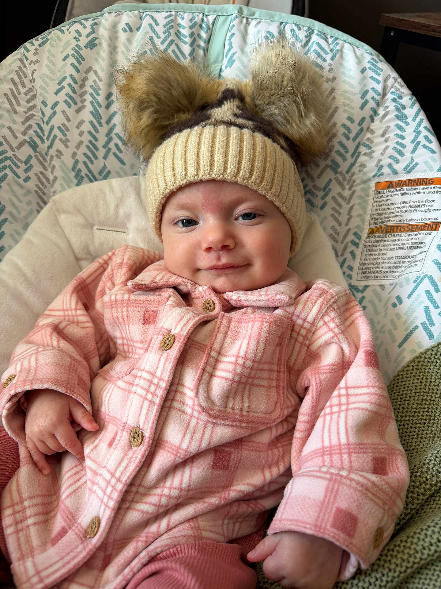 Winsley is registered to the contest to win money with this photo: baby, child, hat, knit_hat, pom_poms, plaid_jacket, pink_clothing, baby_seat, cushion, warning_label, indoor, smiling, face, hand, comfortable, portrait, cozy, apparel, person, warm_clothing
