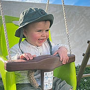 Sacha a rejoint le concours — aidez-le/la à gagner de superbes lots ! baby, child, swing, outdoor, green_swing, hat, bucket_hat, grass, sky, clouds, happy, smiling, playground, grass_with_flowers, white_shirt, pants, socks, daylight, fun, cute