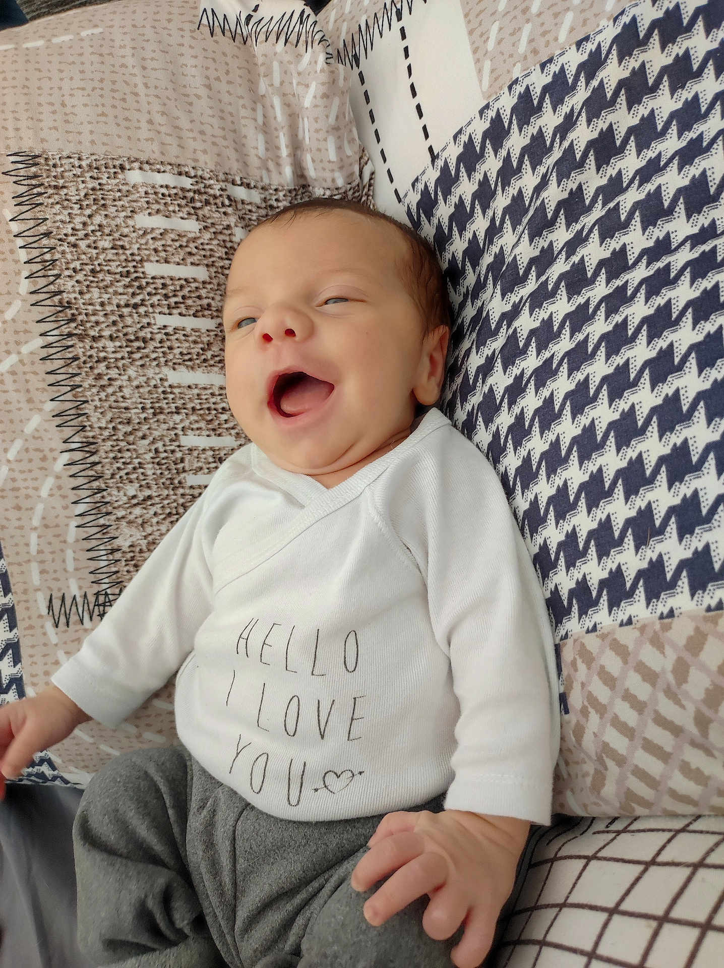 Léandro participe au concours pour gagner de l'argent avec cette photo : baby, infant, smiling, clothing, white_shirt, gray_pants, lying_down, patterned_quilt, happy, face, hand, soft_texture, cute, newborn, young_child, indoors, cozy, expression, person, child
