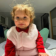 ève a rejoint le concours — aidez-le/la à gagner de superbes lots ! child, toddler, curly_hair, red_clothing, pink_blanket, indoor, bed, blanket, portrait, cute, smiling, person, face, hand, cozy, home, soft_texture, window, furniture, lighting