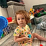 toddler, child, indoor, curly_hair, toy, block, floor, clothing, radiator, furniture, blanket, storage_bin, plastic_toys, wooden_table, colorful, patterned_clothes, sitting, young_child, playtime, home