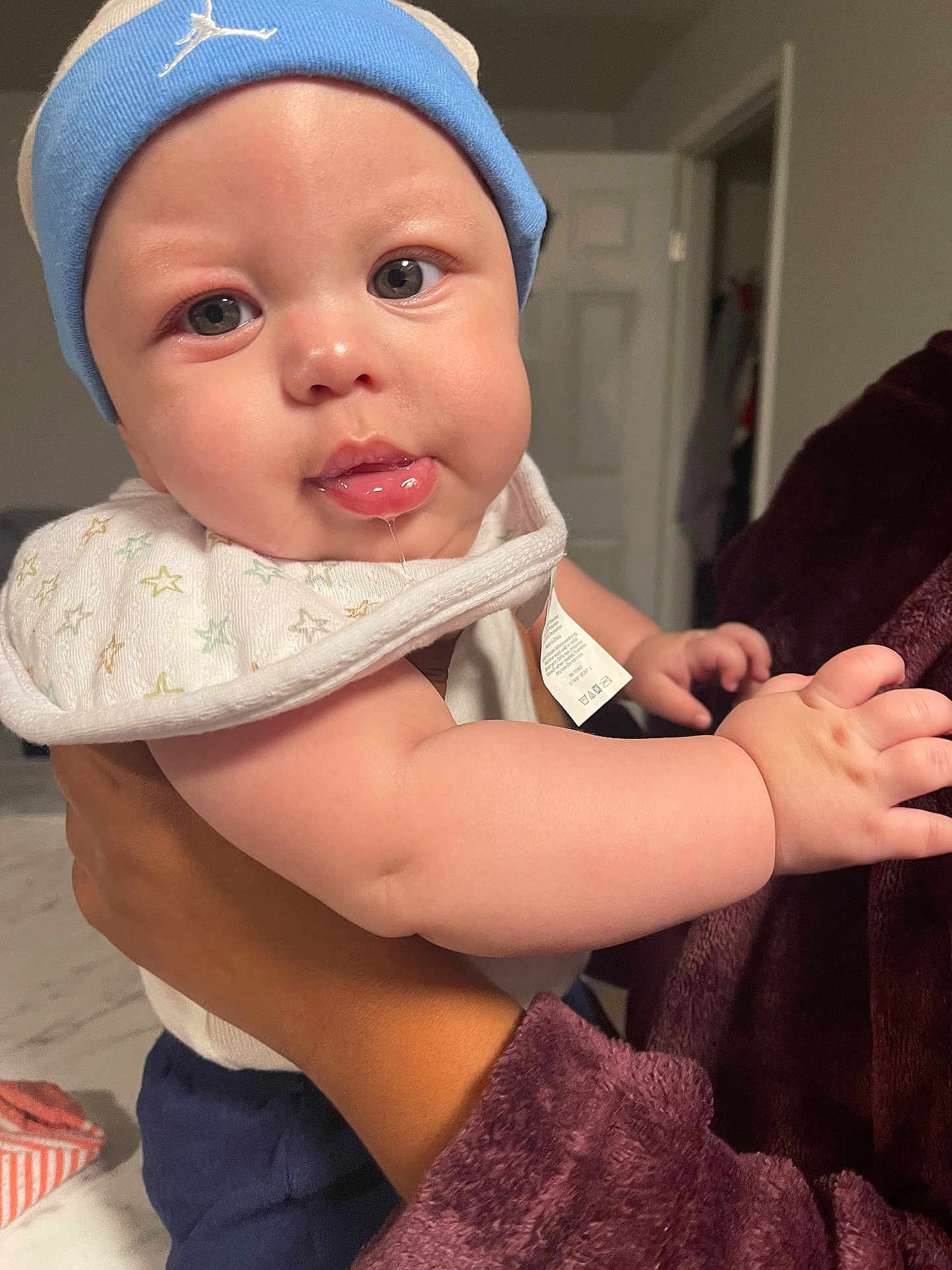 Demilo Johnson is registered to the contest to win money with this photo: baby, drool, bib, blue_beanie, chubby_cheeks, indoor, hand, person, curious, clothing, face, skin, infant, holding, adult_hand, door, fabric, soft_lighting, expression, closeup