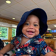 Kameron is registered to the contest to win money with this photo: toddler, child, smiling, hat, bucket_hat, floral_shirt, drool, hand, indoor, restaurant, wooden_furniture, window, ceiling_lights, happy, person, portrait, colorful_clothing, baby, cute, holding