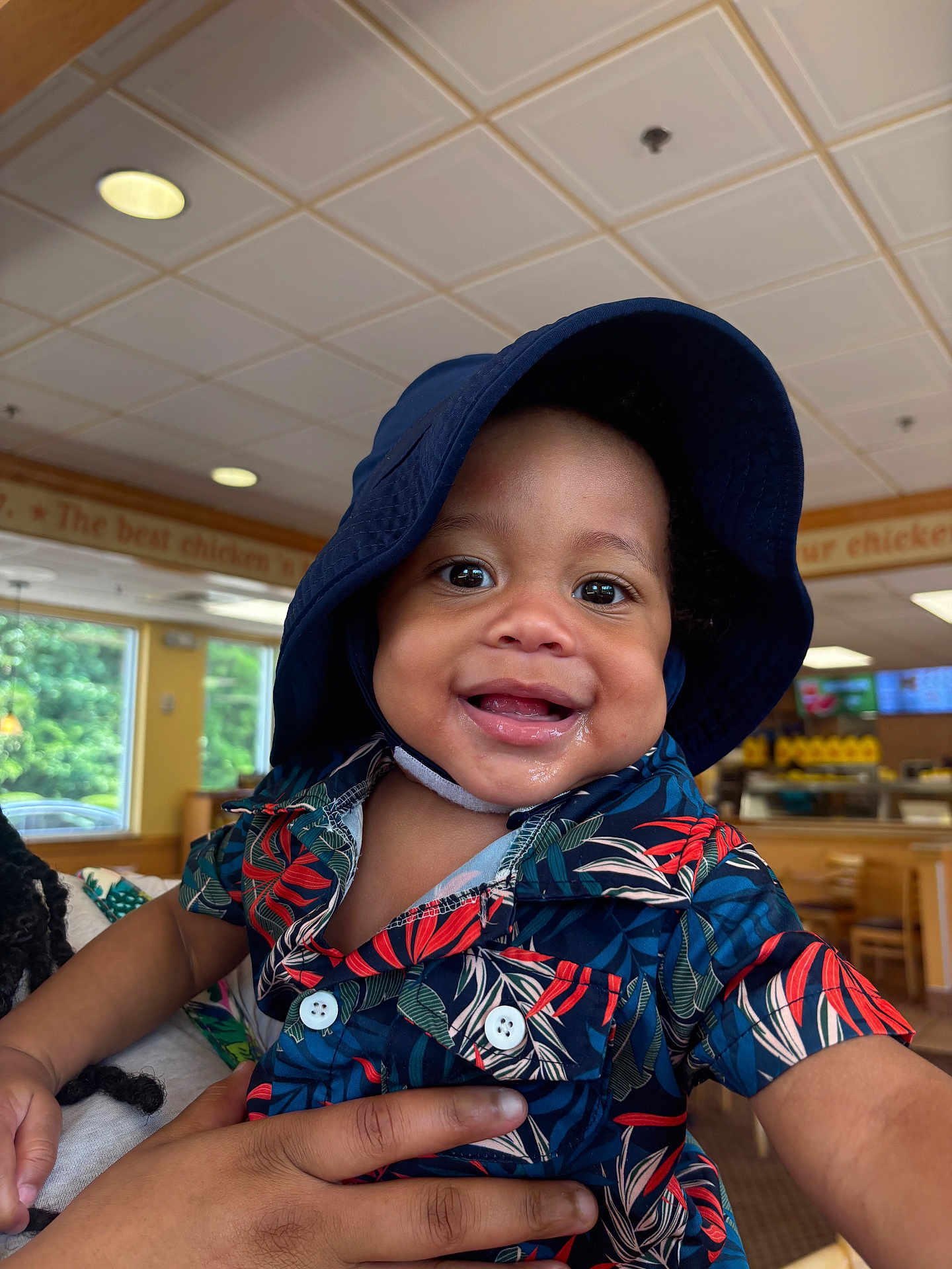 Kameron is registered to the contest to win money with this photo: toddler, child, smiling, hat, bucket_hat, floral_shirt, drool, hand, indoor, restaurant, wooden_furniture, window, ceiling_lights, happy, person, portrait, colorful_clothing, baby, cute, holding