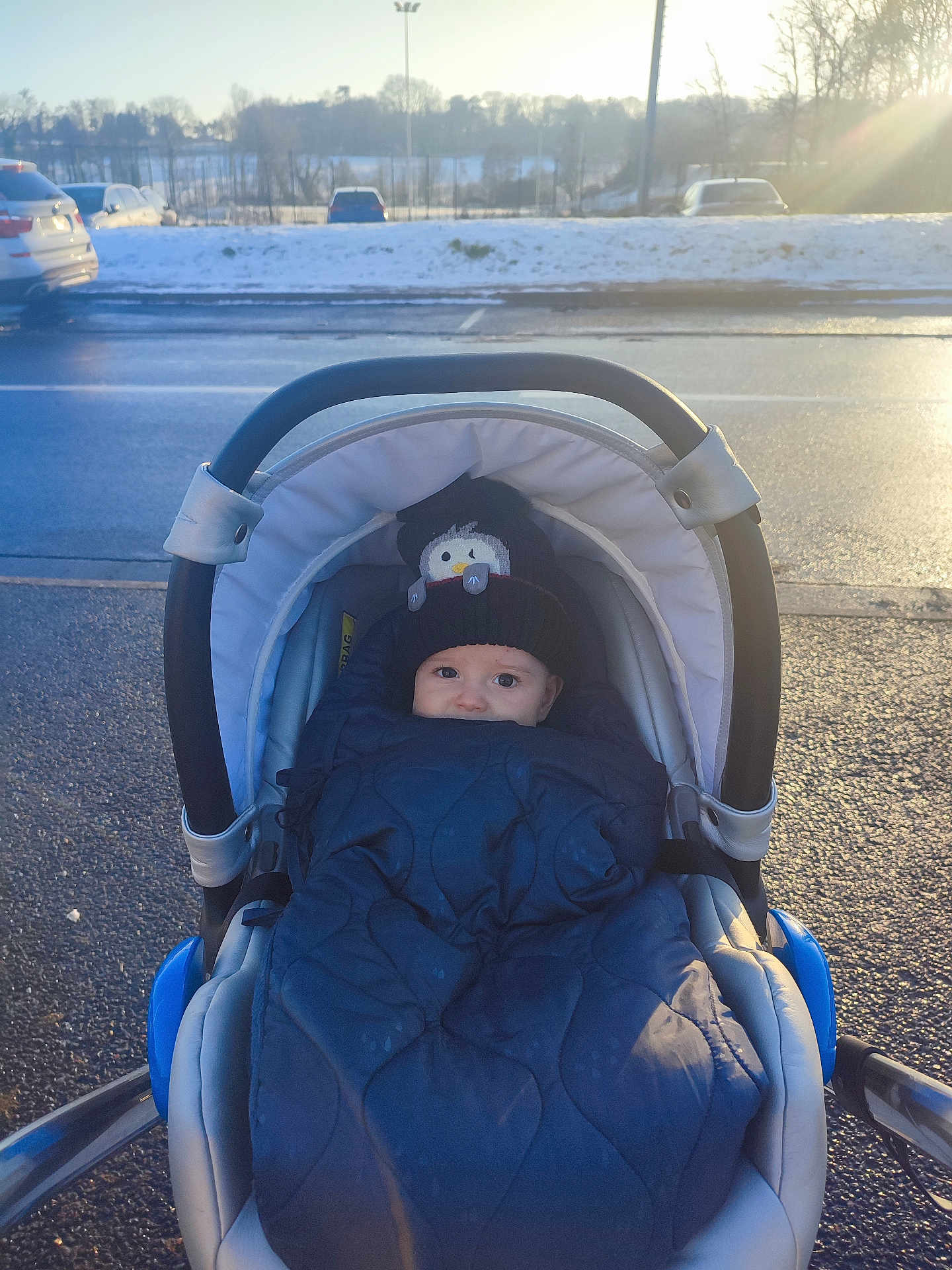Iyed a rejoint le concours — aidez-le/la à gagner de superbes lots ! baby, stroller, blanket, hat, winter, snow, outdoor, child, cold, bundle, parking_lot, car, sunlight, clothing, face, person, daytime, road, warm, cute