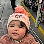 aisle, background, cart, child, clothing, curly_hair, floor, hat, indoor, metal, person, pink, retail, shopping, shopping_cart, store, sweater, toddler, toys, woman