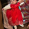 baby, red_dress, headband, blanket, person, patterned_clothing, couch, indoor, infant, soft_texture, comfort, seated, cute, child, holding, footwear, pacifier_clip, colorful, fabric, young_child