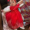 baby, red_dress, headband, bow, blanket, adult, patterned_clothing, seated, infant, cute, face, portrait, sock, pacifier_clip, soft_toy, couch, indoor, person, holding, cozy
