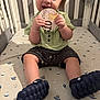 baby, child, crib, card, winnie_the_pooh, green_shirt, shorts, blue_shoes, patterned_sheet, space_theme, sitting, indoor, cute, infant, hands, face, mouth, toy, nursery, cuteness