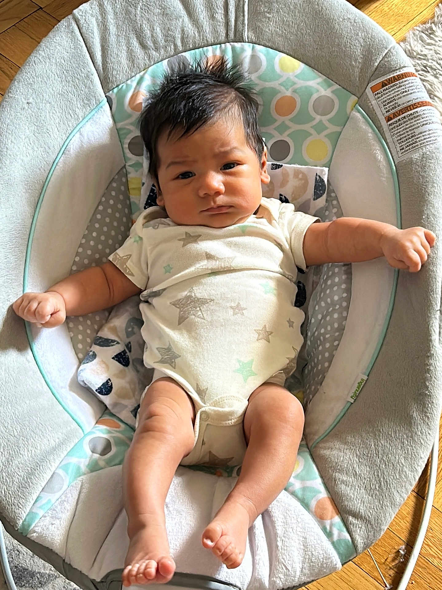 Ethan is registered to the contest to win money with this photo: baby, infant, face, arms, legs, foot, baby_seat, clothing, onesie, pattern, wood_floor, indoor, person, newborn, cute, expression, hair, skin, comfort, child