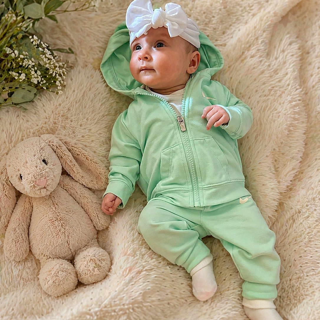 Remi is registered to the contest to win money with this photo: baby, infant, hoodie, headband, white_bow, plush_toy, bunny, blanket, soft_texture, greenery, cute, child, laying_down, cozy, comfortable, indoor, portrait, clothing, socks, adorable
