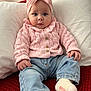baby, child, pink_bow, knitted_sweater, blue_jeans, white_socks, red_blanket, white_pillow, indoor, seated, curious_expression, cute, portrait, infant, cozy_clothing, soft_lighting, casual, hands, face, headwear