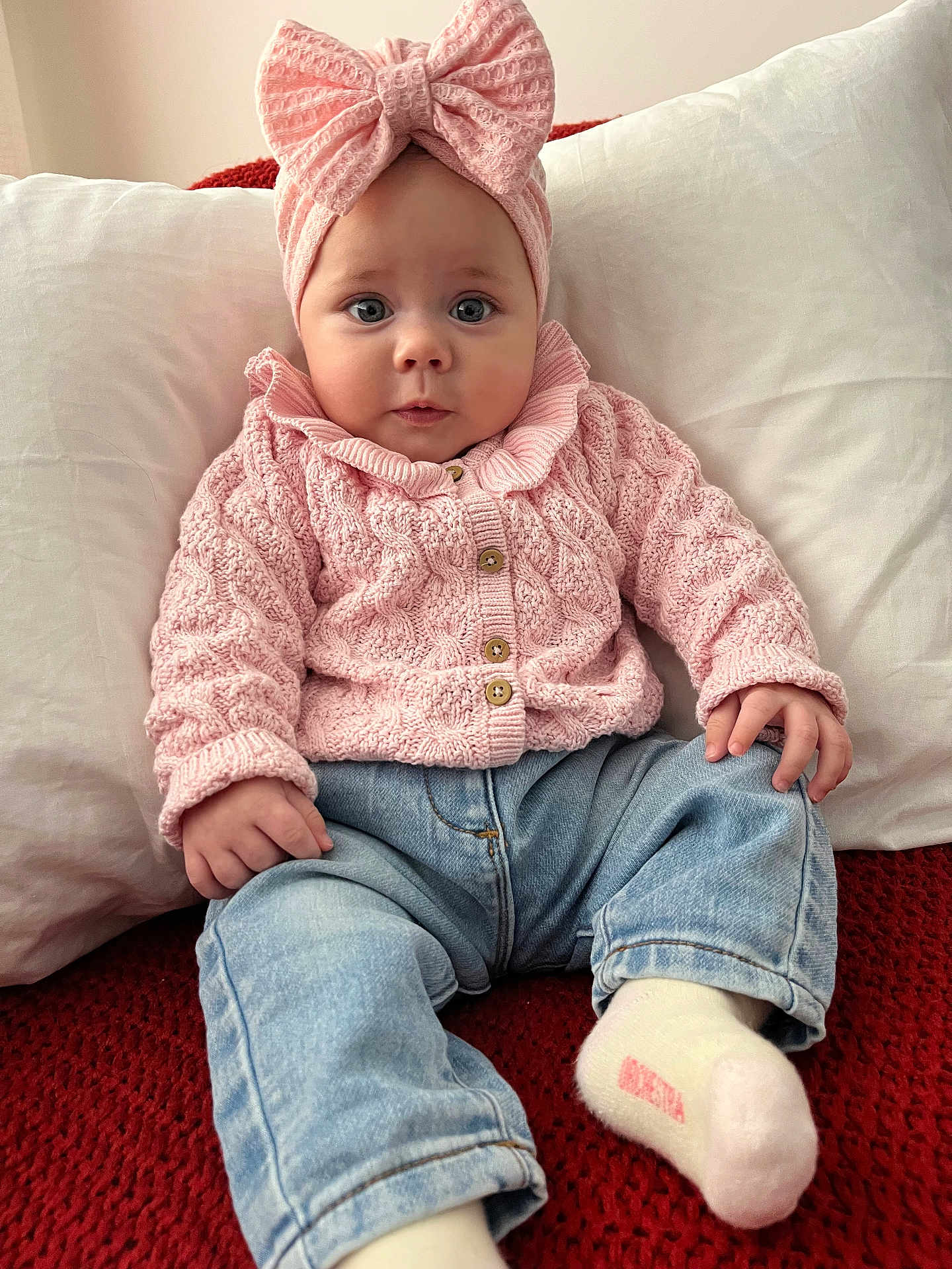 Chloé a rejoint le concours — aidez-le/la à gagner de superbes lots ! baby, child, pink_bow, knitted_sweater, blue_jeans, white_socks, red_blanket, white_pillow, indoor, seated, curious_expression, cute, portrait, infant, cozy_clothing, soft_lighting, casual, hands, face, headwear
