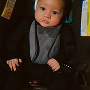 Samir joined the competition — help win amazing prizes! baby, infant, car_seat, black_clothing, bib, white_socks, web_pattern, face, eyes, hands, seatbelt, child, young_child, indoor, portrait, sitting, cute, calm, headwear, person