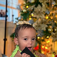 Noah joined the competition — help win amazing prizes! toddler, child, baby, face, hand, hair_bow, black_object, christmas_tree, holiday_lights, decorations, indoors, portrait, cute, curious, festive, smiling, person, holiday, celebration, warm_lighting