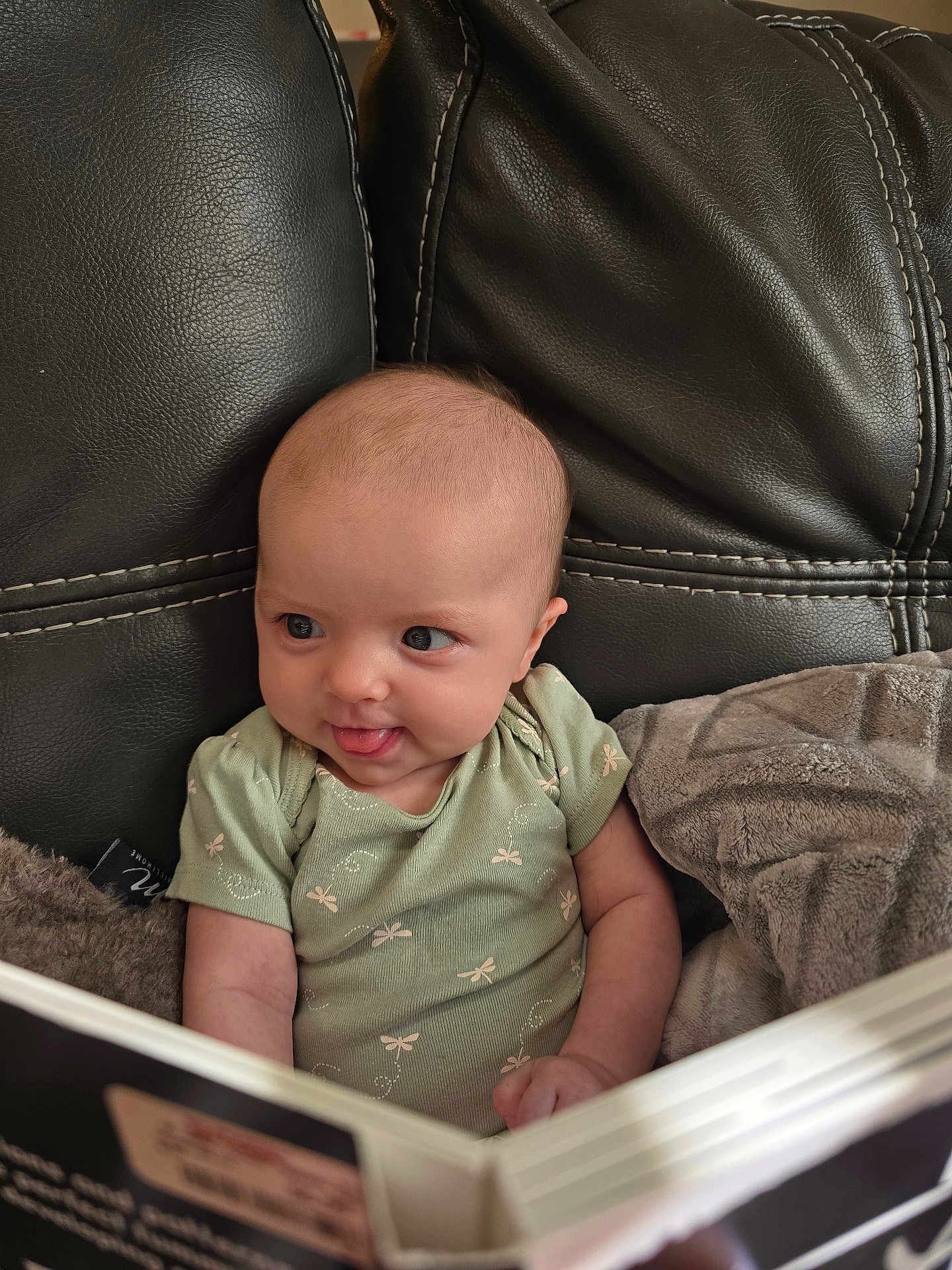 Autum is registered to the contest to win money with this photo: baby, infant, reading, book, couch, leather, blanket, green_clothing, cute, curious, tongue_out, portrait, indoors, child, small_hands, seated, expression, soft_light, closeup, young