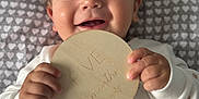 Eric is registered to the contest to win money with this photo: baby, infant, smiling, lying_down, holding, wooden_sign, five_months, blanket, heart_pattern, white_clothing, rainbow, 2025, cute, happy, portrait, child, indoors, soft_lighting, closeup, hands
