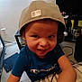 child, boy, smiling, cap, hat, tshirt, champion_brand, indoor, room, floor, carpet, shelf, toy_figurine, electronics, cable, face, portrait, happy, playful, home