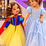 children, girls, princess, costumes, snow_white, blue_dress, yellow_skirt, red_cape, fairy_tale, holding_hands, smiling, curly_hair, hair_bows, indoor, playground, railing, brick_wall, colorful, portrait, dress_up