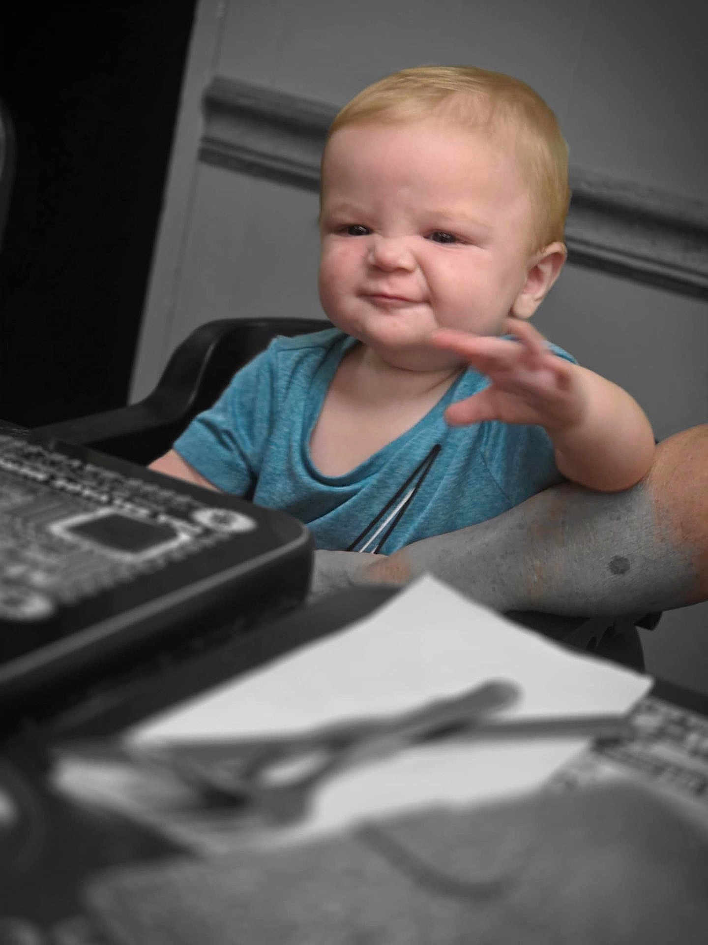 Logan is registered to the contest to win money with this photo: baby, child, blue_shirt, face, expression, hand, arm, table, chair, indoor, person, curly_hair, skin, furniture, focus, blur, background, scrunched_face, reaching, support