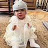 baby, child, helmet, white_dress, socks, carpet, living_room, couch, table, furniture, bow, person, indoor, floor, cute, toddler, headgear, home, carpeted_floor, sitting